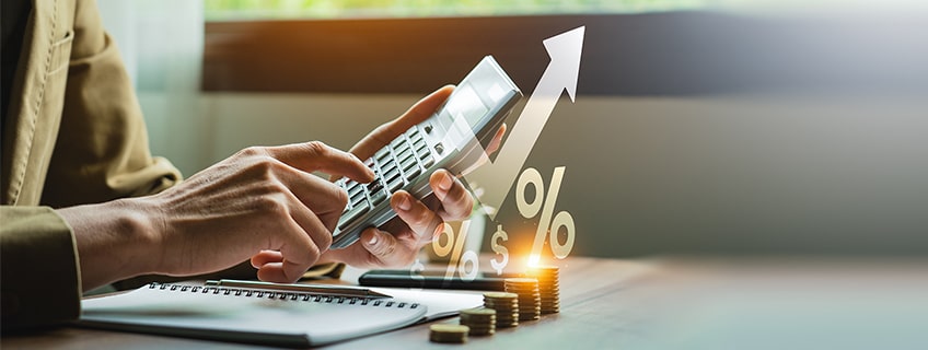 A person calculates returns with coins, a notebook, and rising financial symbols signifying investment comparison. Choose smart with IndusInd Bank Fixed Deposit.
