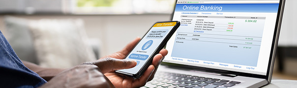 Person managing finances using a phone and laptop, accessing online banking. Open an IndusInd Bank current account to enjoy digital access with low AMB.