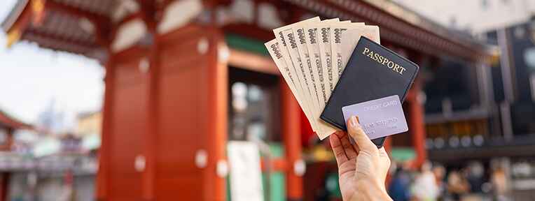 Hand holding cash, passport, and credit card near a landmark. Choose IndusInd Bank Credit Card for ease and savings on travels abroad