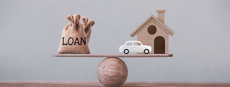 Balancing a bag labeled 'Loan', a car, and a house model on a scale, symbolizing how home improvement loans can enhance your home's appearance and value.